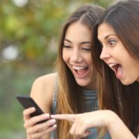 Euphoric friends watching video marketing on a smartphone and pointing at screen surprised